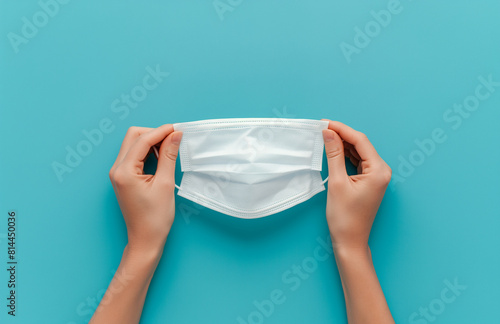Hands holding a white surgical mask against a vibrant teal background, emphasizing health precautions and personal safety during a pandemic - AI generated photo