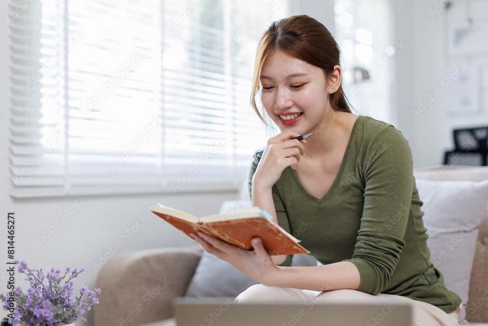 Young asian woman at home sitting on modern chair in front of window relaxing in her living room reading book and drinking coffee or tea Young asian woman at home sitting on modern chair in front of window relaxing in her living room reading book and drinking coffee or tea