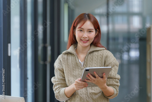 Portrait of a young Asian woman, a company worker in documents smiling and holding a digital tablet, standing over an office background.