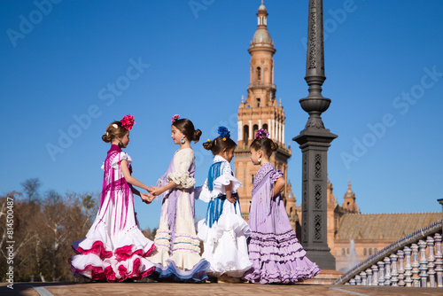 Four little girls dancing flamenco dressed in typical flamenco dress talk to each other in a famous plaza in sevilla, spain. In the background an old square tower.