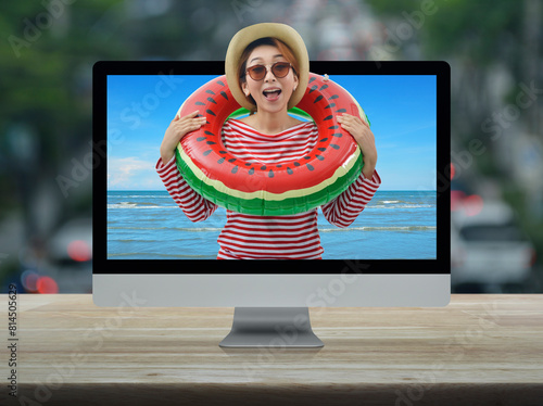 Happy young asian woman wearing red stripped shirt and straw hat in watermelon inflatable ring with tropical sea on computer screen on table over blur of rush hour with cars and road