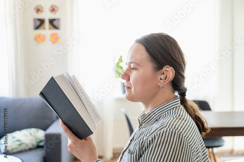 Portrait of young smiling woman with hearing aid on left ear reading book at home