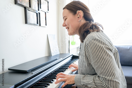 Portrait of young smiling woman with hearing aid on left ear playing piano at home