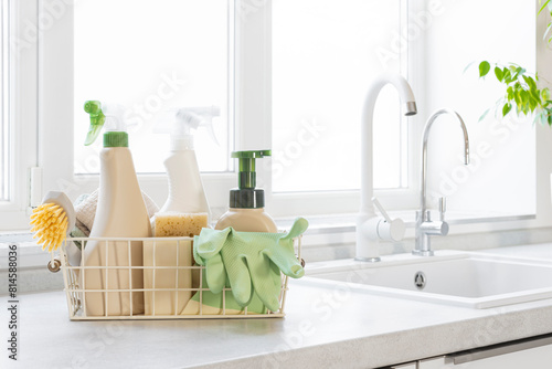 Cleaning products, brushes, sponges, wipes and rubber gloves in basket on kitchen counter near sink and window