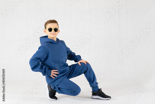 Cheerful boy in a blue tracksuit and sunglasses. Pretty boy,. Baby model. A little boy poses on a white background. Boy posing sitting.Dancing boy