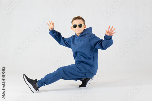 Cheerful boy in a blue tracksuit and sunglasses. Pretty boy,. Baby model. A little boy poses on a white background. Boy posing sitting.Dancing boy