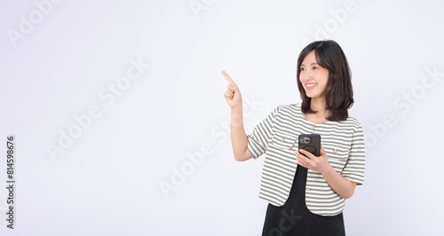 Asian woman is holding a mobile phone and pointing to an empty space on a white background.