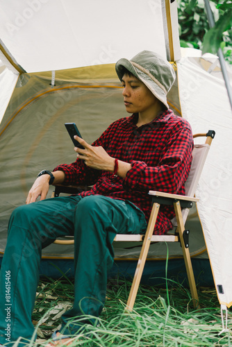 A real transgender Asian lesbian using Surf the web and social media on smartphone smiling in relaxation while sitting outdoors near a tent. © Akira Kaelyn