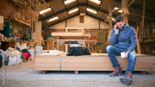 Carpenter Working In Woodwork Workshop With Pet Dog Making Call On Mobile Phone On Coffee Break