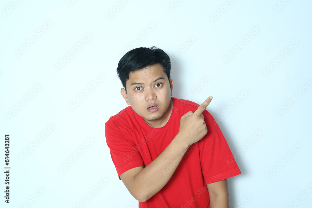 Young Asian man looking camera with worried expression while pointing behind. Wearing a red t-shirt Young Asian man looking camera with worried expression while pointing behind. Wearing a red t-shirt