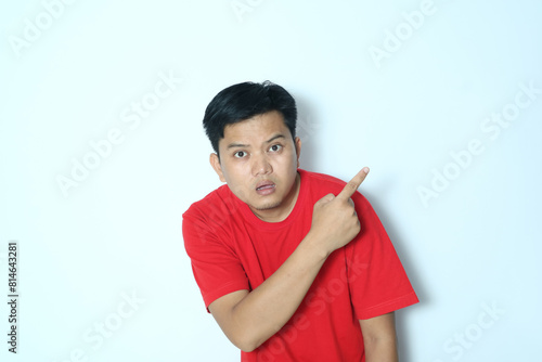 Young Asian man looking camera with worried expression while pointing behind. Wearing a red t-shirt photo