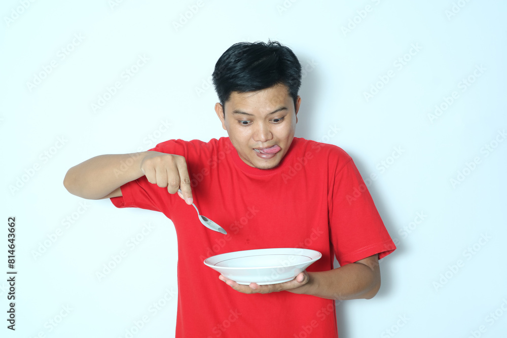 Young Asian man looking to empty eating dinner plate and holding spoon with funny expression. Wearing a red t-shirt Young Asian man looking to empty eating dinner plate and holding spoon with funny expression. Wearing a red t-shirt