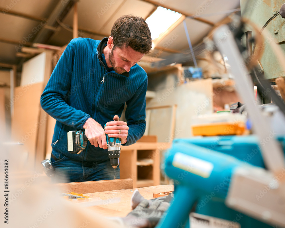Carpenter At Workbench In Workshop Using Cordless Electric Drill