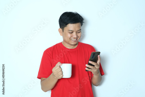 Side view of young Asian man smiling happy looking at a smartphone while holding an white cup. Wearing a red t-shirt photo