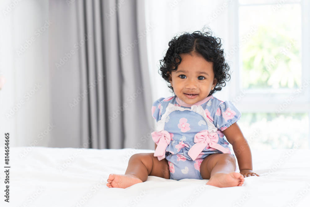 Cute African little newborn 7 months old baby girl wearing clothes with black curly hair sitting on white bed in bedroom at home. Happy adorable small daughter smiling. Copy space Cute African little newborn 7 months old baby girl wearing clothes with black curly hair sitting on white bed in bedroom at home. Happy adorable small daughter smiling. Copy space