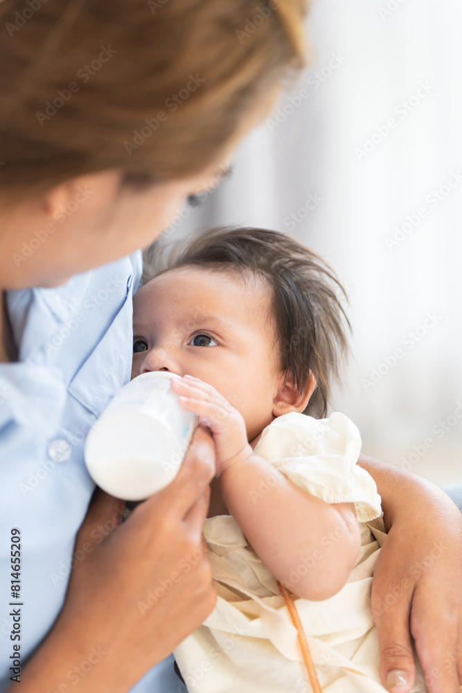 Asian woman feeding milk her cute mixed race little newborn 3 months old baby girl from bottle. Mother holding adorable small daughter in warm arms at home. Copy space