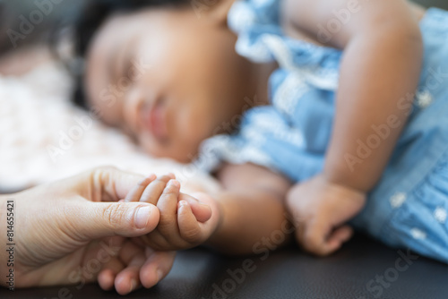 Selective focus on hand of cute African little newborn 7 months old baby girl with black curly hair holding her mother finger while sleeping on sofa with blanket as pillow at home. Family bonding