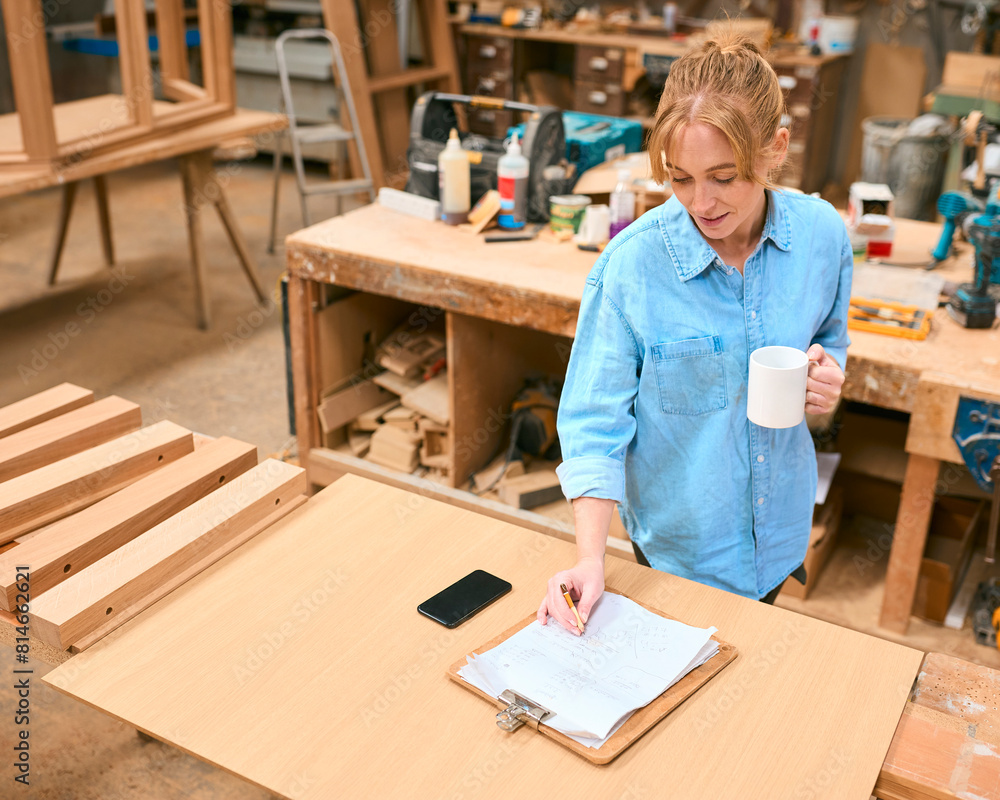 Female Carpenter Working In Woodwork Workshop With Cup Of Coffee Making Notes On Clipboard Female Carpenter Working In Woodwork Workshop With Cup Of Coffee Making Notes On Clipboard