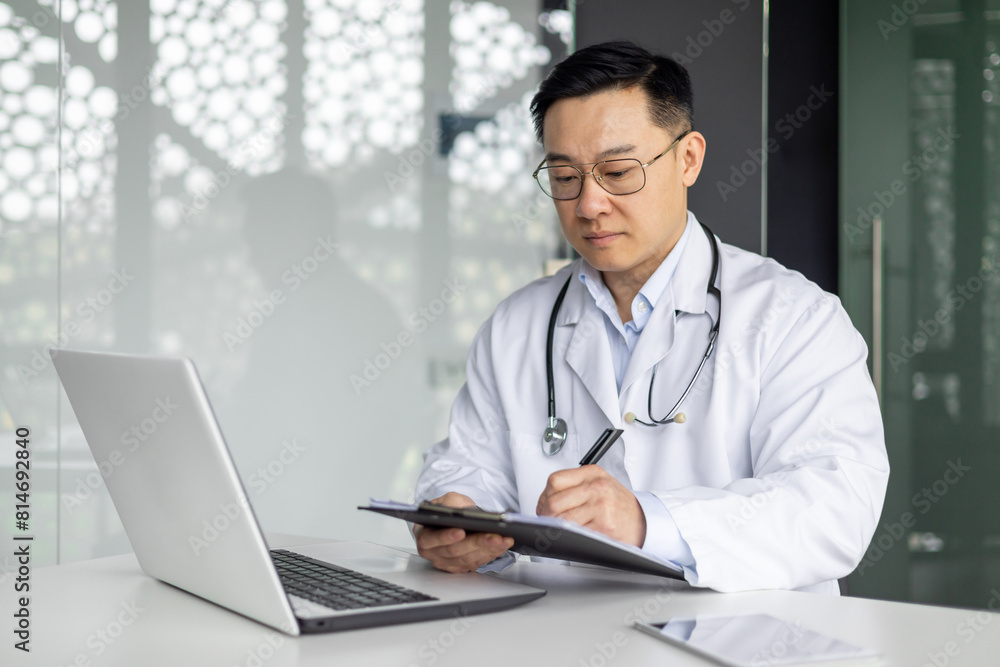 Mature Asian doctor in white coat working on a laptop and writing in a clipboard at the office desk. Professional healthcare and medicine concept. Mature Asian doctor in white coat working on a laptop and writing in a clipboard at the office desk. Professional healthcare and medicine concept.