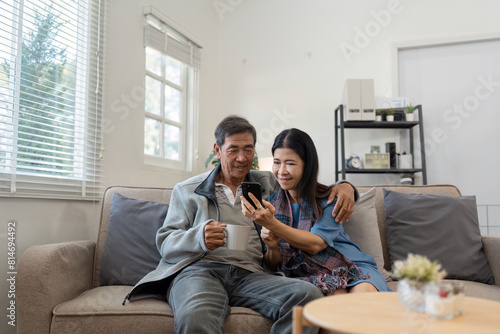 Happy married retirement couple looking at mobile using smartphone mobile technology device together, relaxing on couch doing online shopping