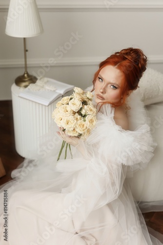 A beautiful lady with fiery red hair elegantly holding a bunch of white roses in a white attire indoors