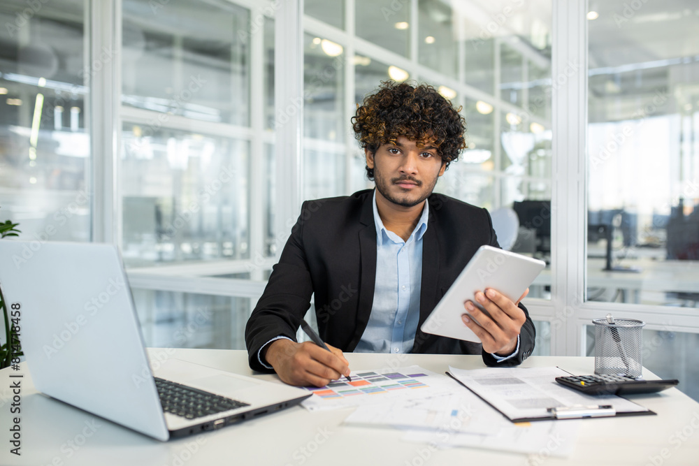 Confident young businessman using a tablet and working on documents in a modern office setting with laptop and papers Confident young businessman using a tablet and working on documents in a modern office setting with laptop and papers