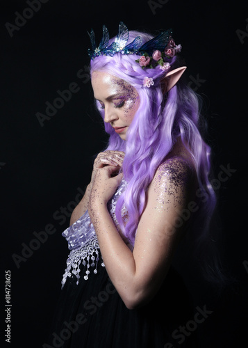 artistic portrait of beautiful female model with long purple hair wearing a fantasy fairy crown and elf ears,  gestural  pose with hands reaching up to touch face.  isolated on dark studio background.