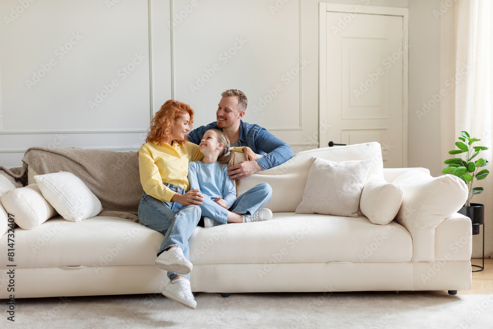 Cheerful family of three sitting on couch in light living room, parent and their daughter enjoy time together at home. Parenthood happiness
