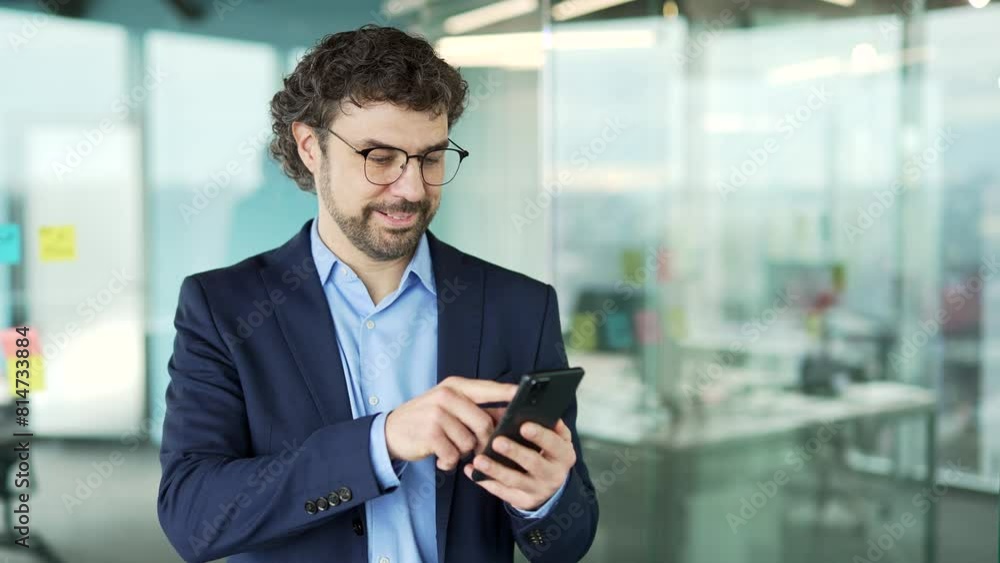 Confident businessman in a suit is using phone standing at workplace in business office. Man worker works on application, typing a message, chatting with a client, banking, ordering or browsing online Confident businessman in a suit is using phone standing at workplace in business office. Man worker works on application, typing a message, chatting with a client, banking, ordering or browsing online