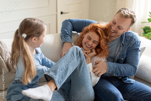 Obraz na płótnie Joyful family of three having fun at home on sofa, man and girl tickling mother