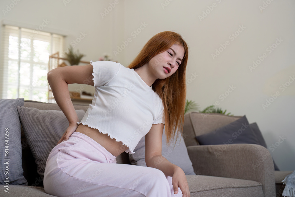 Asian woman having backache and touching her back in living room at home Asian woman having backache and touching her back in living room at home