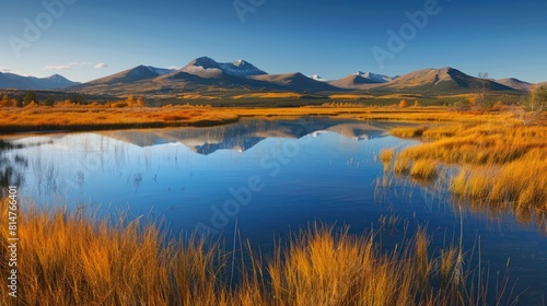 The image showcases a tranquil mountain range reflected in a calm lake surrounded by golden grasses under a clear blue sky