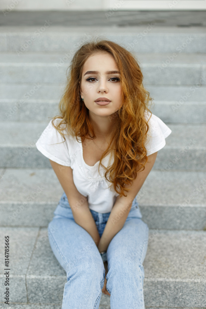 beautiful sexy fashionable woman with red hair in fashion casual clothes with jeans sits on the steps and looks at the camera beautiful sexy fashionable woman with red hair in fashion casual clothes with jeans sits on the steps and looks at the camera