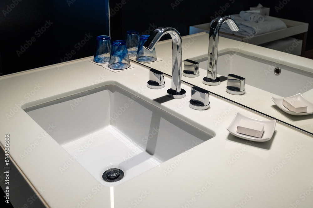 A clean ceramic water sink with a rounded rectangular basin, the faucet has hot and cold handles, soap dish holding a bar of soap, all in a hotel bathroom. In the mirror reflection are folded towels.