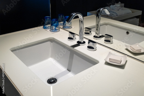 A clean ceramic water sink with a rounded rectangular basin  the faucet has hot and cold handles  soap dish holding a bar of soap  all in a hotel bathroom. In the mirror reflection are folded towels.