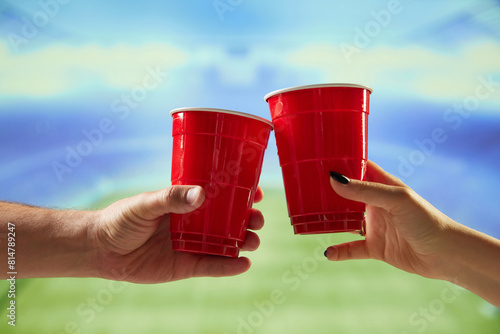 Hands holding red mugs with beer and clicking with blurred sport open air stadium on background. Concept of game, leisure, championship, tournament, emotions