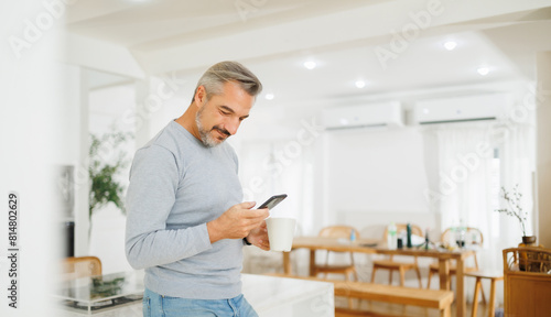 Smiling mature man with grey hair enjoying a cup of coffee and smartphone in a cozy kitchen setting