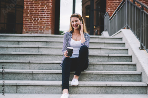 Smiling and happy young woman speaking per modern cellphone and holding cup of coffee while being in big city and sitting at stairs with building on background, concept of technology and communication © BullRun