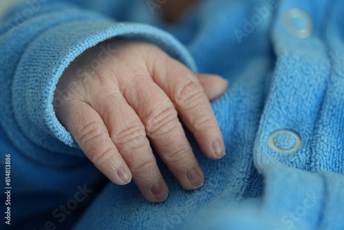 A macro shot of a newborn baby's tiny hand with fingers gently curled, dressed in a blue onesie, epitomizing the fragility and beauty of new life. © Marco