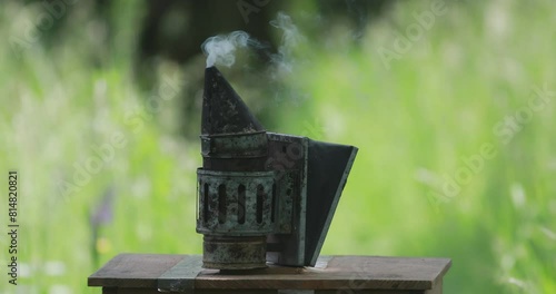 An old bee smoker placed on the top of a beehive lid puffing smoke in the air  photo