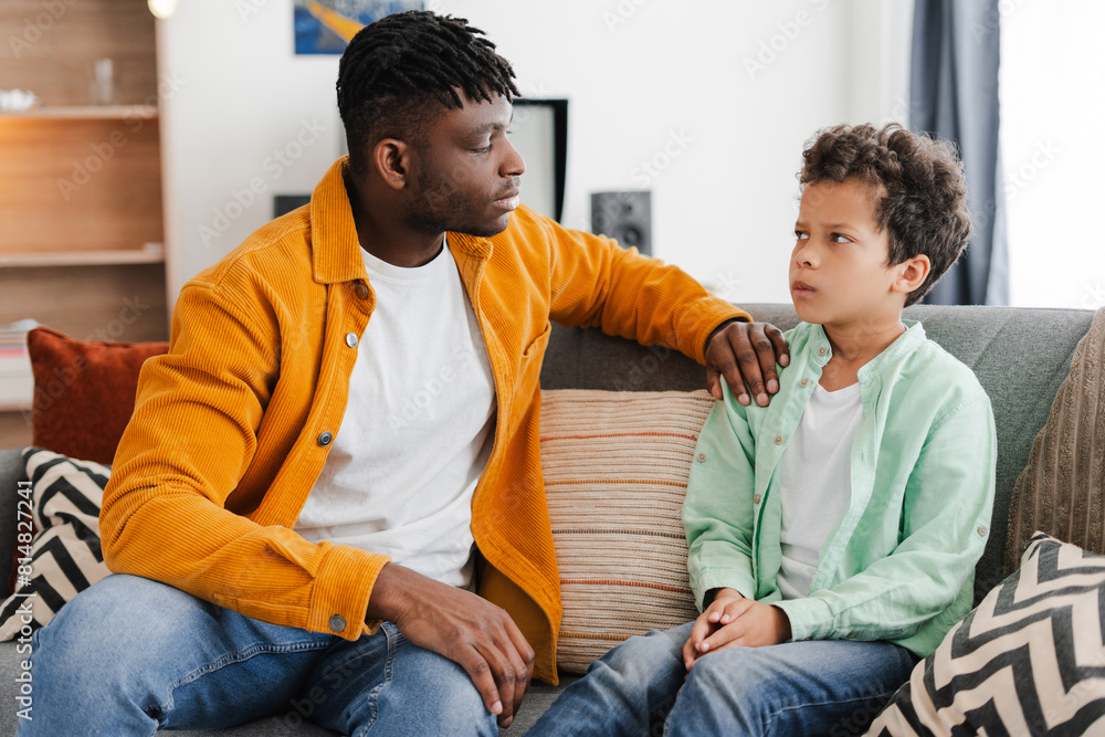 Young African American man talking with with little offended son while sitting on comfortable sofa Young African American man talking with with little offended son while sitting on comfortable sofa