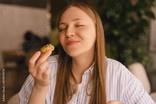 Young blonde woman eating eclair sitting in cafe. Girl bite piece of croissant look joyful at restaurant. Cheat meal day concept. Woman is preparing with appetite to eat eclair. Enjoy pistachio bakery