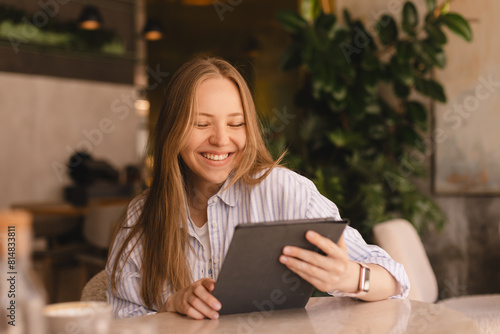 Attractive happy smiling blonde woman wear shirt sitting with tablet computer in cafe. Beauty girl student using tablet computer for social media, studying or reading news.