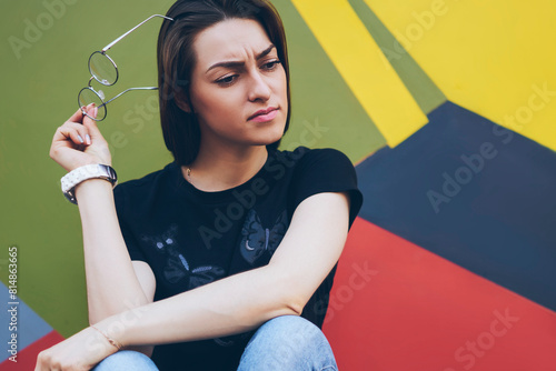Young pondering hipster girl feeling puzzled while thinking about what to do on weekend, pensive woman with spectacles sitting on colorful background, confusion face and expression concept photo