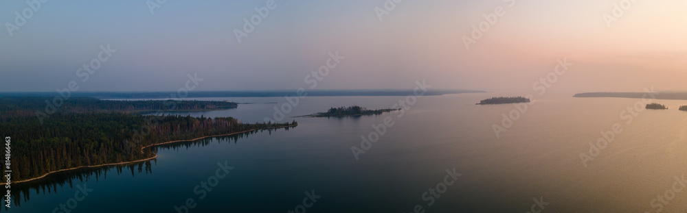 Aerial panorama of a large lake and surrounding forest as the sun rises on the right side of the picture. The sky varies in color from pale blue to orange. The calm water reflects the colors.
