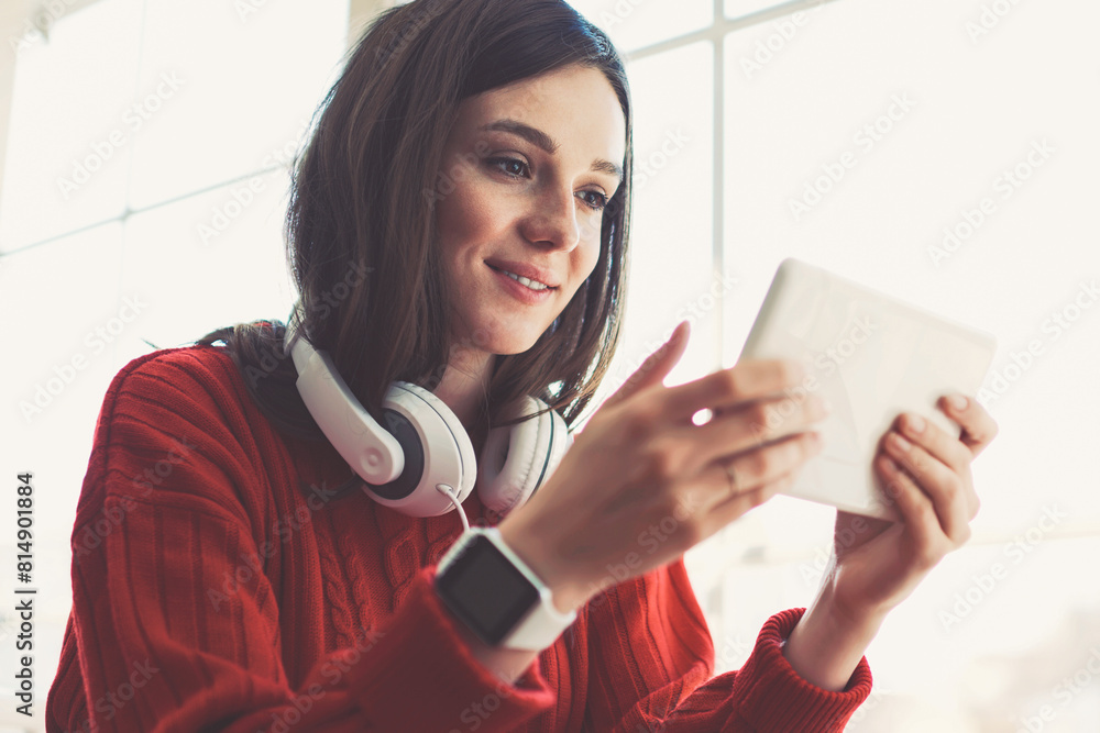 Naklejka premium Caucasian hipster girl enjoying time at coffee shop watching video online via portable pc while waiting friend, young smiling female gamer student passing next level via digital tablet app on leisure