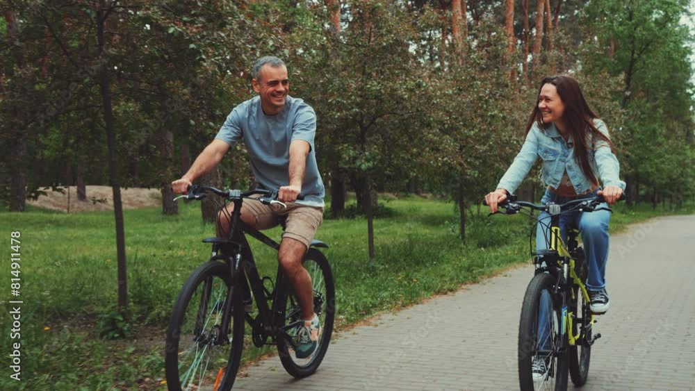 Smiling couple rides bicycles together along park path. Man and woman share joyful moment, cycling side by side surrounded by lush greenery and trees. Outdoor activity promotes healthy lifestyle Smiling couple rides bicycles together along park path. Man and woman share joyful moment, cycling side by side surrounded by lush greenery and trees. Outdoor activity promotes healthy lifestyle