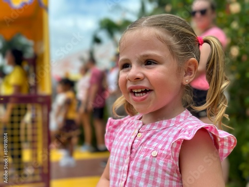 A joyful girl in a pink gingham dress smiles broadly at an amusement park, her ponytail swaying, with colorful rides and people in the background. © David