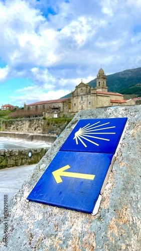 Blue sign with shell and arrow of the Portuguese Camino de Santiago, along the coast, with the sea and the Monastery of Oia
