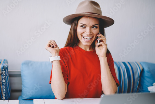 Cheerful female teen communicated with friend via smartphone device sitting at cafeteria table with modern computer, excited woman in trendy titfer looking away and laughing during telephone call photo